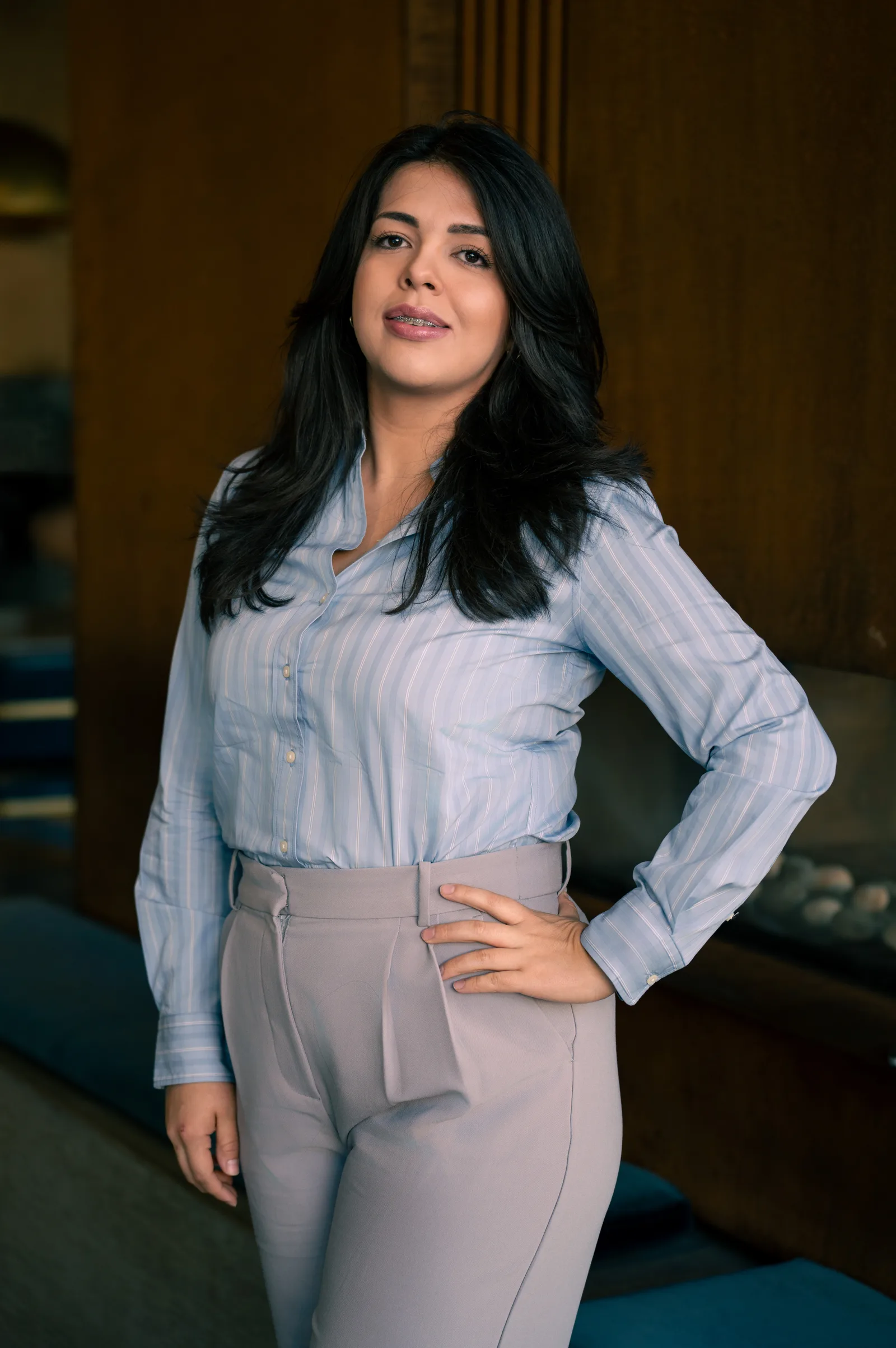 Portrait of Ruby Medina in a light blue shirt, hand on hip, in a warm wood-panelled room